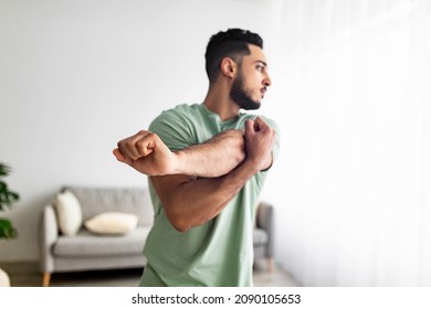Athletic young Arab guy stretching arm muscles, doing warmup exercises at home, selective focus. Millennial middle Eastern man having domestic training, working out indoors