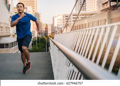 Athletic Young Asian Man In Sportswear Looking Focused Crossing Over A Bridge While Out For A Run Through The City