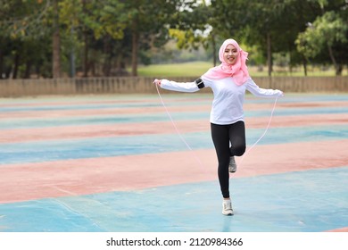 Athletic Young Asian Woman In Muslim Sportswear Standing And Skipping Rope Outdoor For Morning Exercise. Active Girl Work Out On Biceps Exercise With Green Tree Background. Sport Concept