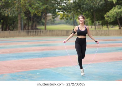 Athletic Young Asian Woman In Sportswear Standing And Skipping Rope Outdoor For Morning Exercise. Active And Healthy Girl Workout On Biceps Exercise With Tree Background. Sport And Lifestyle Concept