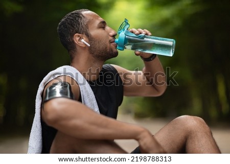 Athletic young black man drinking water while having break, training at public park, using wireless earbuds, listening to music while exercising, copy space. Hydration and sport concept