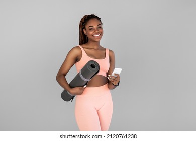 Athletic young black woman in sportswear holding fitness mat and smartphone on grey studio background. Millennial African American female ready for online yoga or pilates class - Powered by Shutterstock