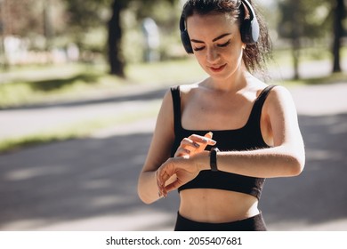 Athletic Young Happy Woman Runing With Headphones In The Park, The Girl Looks At The Watch Outdoor Workout. Sport And Healthy Active Lifestyle Concept. Black Clothing For Sports