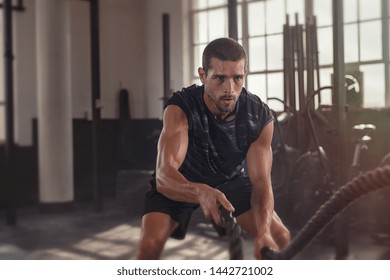 Athletic young man doing some fitness exercises with a rope. Determined fit guy doing battle ropes exercise at the grunge gym. Handsome man training with effort.