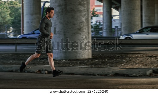 Athletic Young Man in Earphones and\
Sports Outfit is Jogging in the Street. He is Running in an Urban\
Environment Under a bridge with Cars in the\
Background.