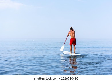 Athletic young man paddling on a sup board on the quiet sea - Stand up paddle boarder training on a rowing board on a flat calm sea - Back view and copy space for text