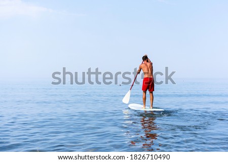 Athletic young man paddling on a sup board on the quiet sea - Stand up paddle boarder training on a rowing board on a flat calm sea - Back view and copy space for text