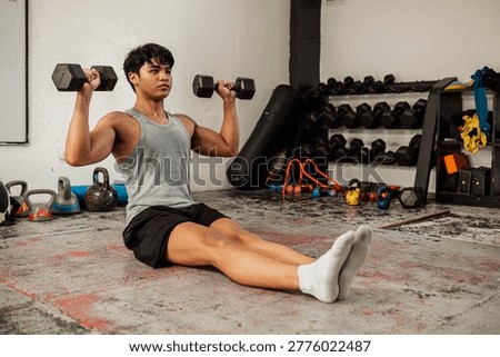 Athletic young man performs seated dumbbell Z press on gym floor, building shoulder strength and core stability in hardcore training space.
