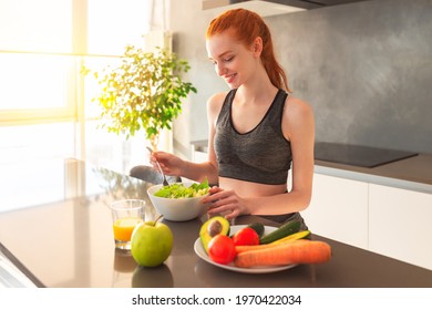 Athletic young red haired woman in the home kitchen eating a healthy salad - Shutterstock ID 1970422034