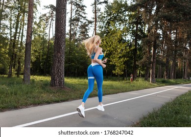 Athletic young woman jogging in fashionable blue sportswear in gym shoes in public park against bright sunset. Sporty girl blonde with gorgeous body with sexy ass running through forest. Back view .