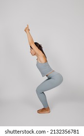 Athletic Young Woman Posing. Concept Of Healthy Lifestyle, Brunette In Fitness Suit Stands In Yoga Pose On Isolated Gray Background, She Bent Her Knees And Raised Her Arms Up, Side View. 