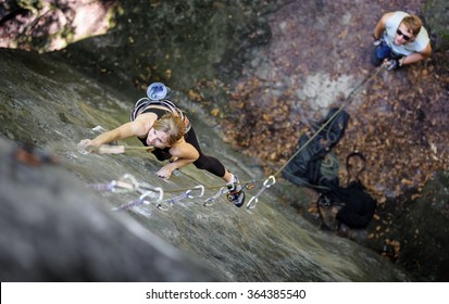 Athletic young woman rock climbing with carbines and rope on summer day. Man standing on the ground insuring the climber