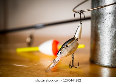 ATLANTA, GEORGIA - DECEMBER 29, 2018 : Fishing Lure Hanging From Metal Minnow Bucket Handle. Bass Fishing Tackle Including Crank Bait Lure, Fishing Rod And Bobber. White And Wood Background.