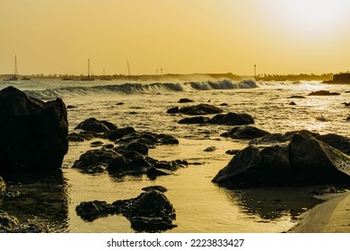 The Atlantic ocean with golden sunset sky above against the backdrop of the sailboats and rocks on the beach coast in foreground in Santa Maria, Sal island, Cape Verde.