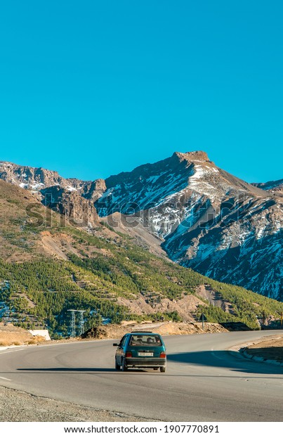 Atlas Mountains,\
Morocco - January 18, 2021 - vertical view of a Fiat driving on a\
road in the High Atlas Mountains in Morocco with snow-capped peaks\
in the background