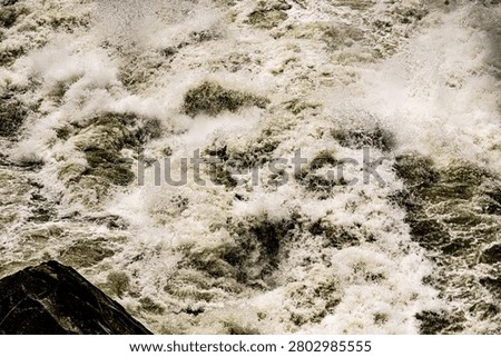 An atmospheric and dramatic view of rushing river rapids, featuring intense white foam and splashing water swirling around large, dark rock formations. The play of light and shadow on the water surface emphasizes the sheer intensity and speed of the current. A compelling image representing the unstoppable force of nature and river landscapes.