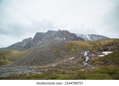 Atmospheric Landscape With Clear Mountain Stream From Hills Against Snowy Mountain Range With Peaked Top In Low Clouds. Beautiful Mountain Creek And High Sharp Rocks Under Cloudy Sky In Gloomy Weather