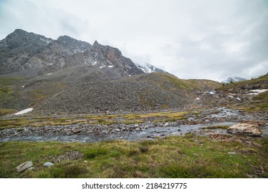 Atmospheric Landscape With Clear Mountain Stream From Hills Against Snowy Mountain Range With Peaked Top In Low Clouds. Beautiful Mountain Creek And High Sharp Rocks Under Cloudy Sky In Gloomy Weather