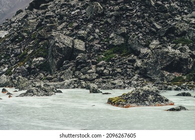 Atmospheric landscape with mountain creek among moraines in rainy weather. Bleak scenery with milky river among rocks. Gloomy view to mountain river. Stones with moss and lichen in milk water stream.