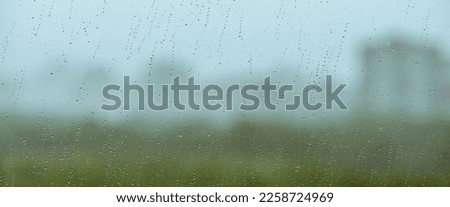 Atmospheric minimal backdrop with rain droplets on glass. Wet window with rainy drops and dirt spots closeup. Blurry buildings and green trees under blue sky against dirty window glass with raindrops.