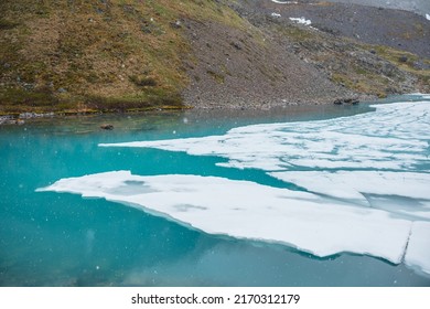 Atmospheric mountain landscape with frozen alpine lake during snowfall. Clear ice floats on water surface of lake. Awesome mountain scenery with icy mountain lake. Flakes of snow on water background.