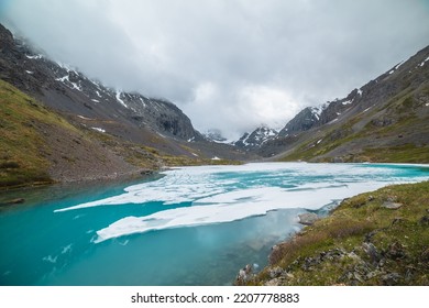 Atmospheric Mountain Landscape With Frozen Alpine Lake And High Snowy Mountains. Awesome Overcast Scenery With Icy Mountain Lake On Background Of Snow Mountains In Low Clouds. Scenic View To Ice Lake.