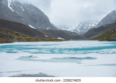 Atmospheric Mountain Landscape With Frozen Alpine Lake And High Snowy Mountains. Awesome Overcast Scenery With Icy Mountain Lake On Background Of Snow Mountains In Low Clouds. Scenic View To Ice Lake.