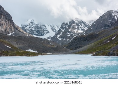 Atmospheric Mountain Landscape With Frozen Alpine Lake And High Snowy Mountains. Awesome Overcast Scenery With Icy Mountain Lake On Background Of Snow Mountains In Low Clouds. Scenic View To Ice Lake.