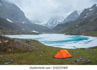 Atmospheric mountain landscape with orange tent near frozen alpine lake during snowfall. Awesome overcast scenery with snowfall in highland valley with ice mountain lake with view to snow mountains.