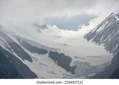 Atmospheric mountain landscape with silhouettes of sharp rocks and sunlit glacier in low clouds at changeable weather. Dramatic view to large glacier tongue in snow mountains in sunlight in low clouds