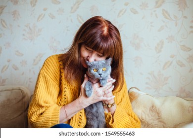 Atmospheric Portrait Of Mature Red Hair Woman In Yellow Sweater Kissing Gray Cat While Sitting On Sofa At Cozy Home Interior. Friendship Pet Lover Concept.