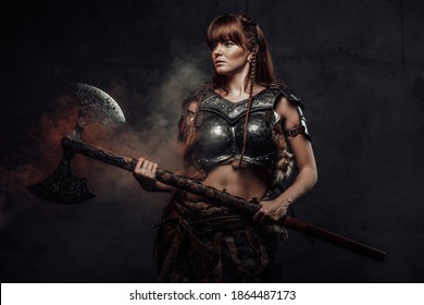 Atmospheric portrait of scandinavian amazon with brown hairs dressed in light armour holding two handed axe in dark background.