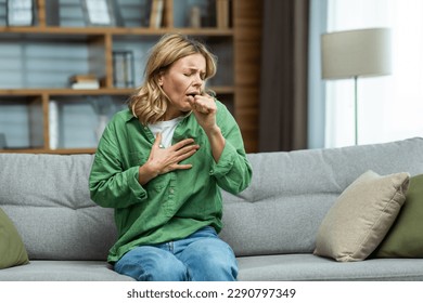 An attack of asthma, allergies at home. An elderly woman is sitting on the sofa, holding her chest and coughing, feeling severe pain, suffocating.
