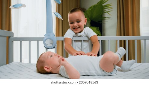 Attaractive playful older brother standing near the crib smilling and playing with the newborn. Concept of raising children and happy family. - Powered by Shutterstock
