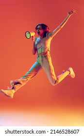 Attention. One slim beautiful excited girl jumping with megaphone isolated on peach color background in neon light, filter. Concept of emotions, expression, youth, aspiration, sales. Copy space for ad