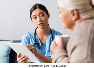 Attentive Asian Nurse Looking At Senior Woman While Holding Digital Tablet