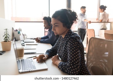 Attentive Call Centre Indian Woman Employee Sitting In Modern Shared Office Working Using Computer Wearing Headset With Microphone Answering Or Making Telephone Calls, Give Support To Company Clients