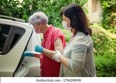 An attentive caregiver helps her elderly client to get into a car to travel for regular medical appointments. Both wear protective masks.