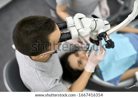 Attentive dentist in a gray uniform with protective mask and white latex gloves looks in a dental microscope while treats his female patient in a blue bib in a clinic. Top view photo. Horizontal.