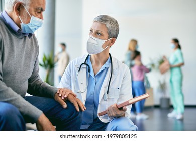 Attentive Doctor Consoling Her Senior Patient And Communicating With Him In Waiting Room At The Hospital While Wearing Protective Face Mask Due To COVID-19 Pandemic. 