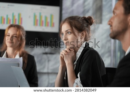 Attentive faces during a briefing: bun-haired businesswoman centred and focused, bar charts colourful on the screen behind her ginger teammate in a bright modern coworking office.