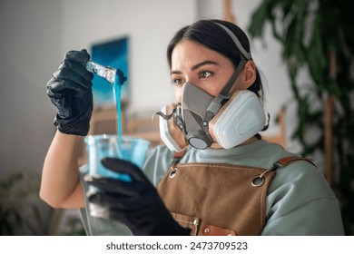 Attentive female artist wearing respirator and latex gloves tests blue epoxy resin composition for creative working in art studio. Focused female artist examines liquid material for seascape paintings - Powered by Shutterstock