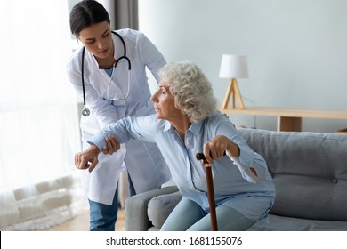 Attentive Female Nurse Give Help Disabled Senior Woman With Walking Stick Stand Up From Couch At Home, Caring Young Caregiver Or Doctor Assist Sick Old Lady With Cane, Elderly Healthcare Concept