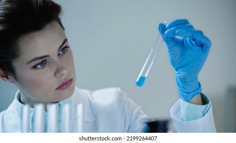 Attentive Female Scientist Looking At Blue Liquid In Test Tube And Writing Down Results