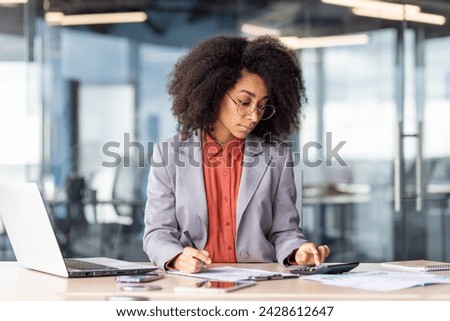 Attentive financial manager checking accuracy of counting on calculator and analyzing bills laying on desktop with laptop. Concentrated female in suit managing expenses of company in modern office.