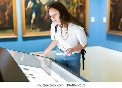 Attentive Girl Visitor At The Museum Looks At The Exposition Behind A Glass Display Case, Holding An Information ..booklet