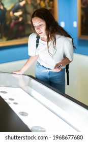 Attentive Girl Visitor At The Museum Looks At The Exposition Behind A Glass Display Case, Holding An Information ..booklet