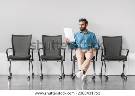 Attentive indian male applicant scrutinizing document while sitting in interview waiting room, symbolizing preparation and focus for potential job opportunity