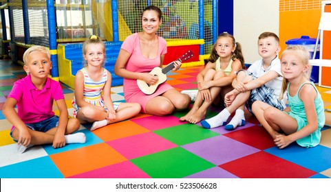 Attentive Kids Sitting Around Attractive Teacher With Small Guitar And Listening To Music In Class