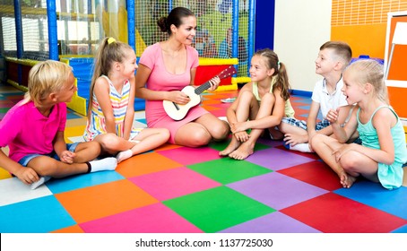 Attentive Kids Sitting Around Cheerful Teacher With Small Guitar And Listening To Music In Class